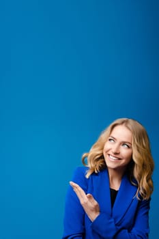 Beautiful caucasian woman with light wavy hair in a blue jacket likes it isolated on blue background