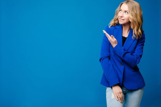 Stock photo portrait of a smiling gorgeous blonde Caucasian woman in blue jacket showing at something with her hand. Copy space or blank space on bright blue background.