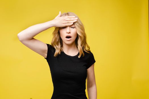 Stock photo of a blonde Caucasian woman in black t-shirt holding hand on forehead with open mouth and eyes closed. Demonstrating headache. Woman suffering from headache. Yellow background.