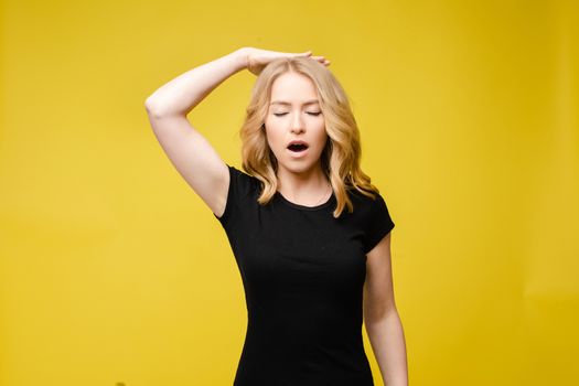 Stock photo portrait of a young Caucasian woman with blonde hair wearing black t-shirt. Woman with eyes closed and open mouth demonstrating awful migraine holding hand on the back of her head. Isolate on yellow background.