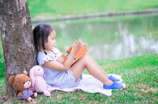 cute little girl  reading a book with a doll in the park