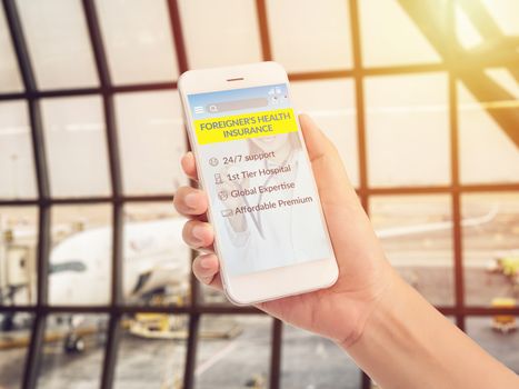 hand holding mobile phone with advertising screen for foreigner's health insurance application with good service and covered benefit condition, airport terminal with airplane parking at background