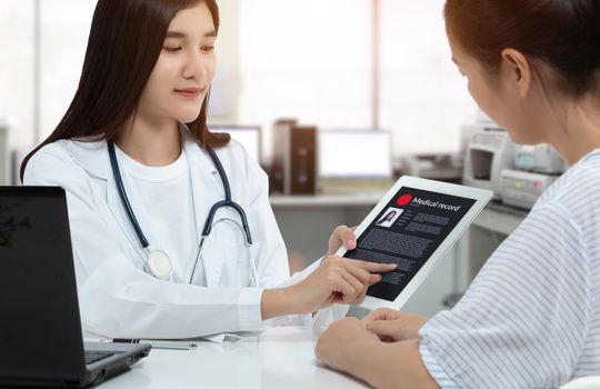 Asian woman doctor holding tablet with patient medical record on screen while consulting patient in medical consultation room.