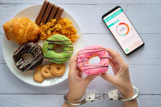 Calories control, food diet and weight loss concept. top view of two hands was control by handcuff, holding doughnut with Calorie counter application on smartphone on dining table