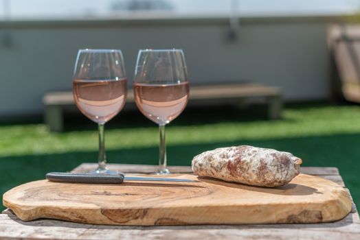 Two glasses of rose wine and a dried sausage with a knife on a wooden table in a garden on a sunny day