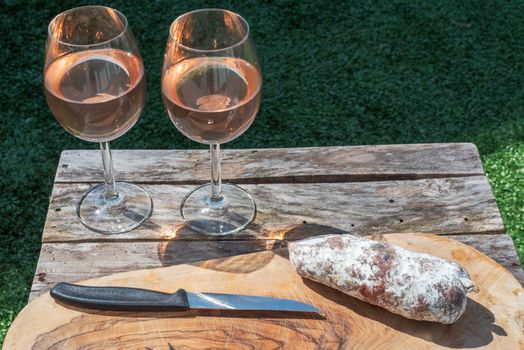 Two glasses of rose wine and a dried sausage with a knife on a wooden table in a garden on a sunny day