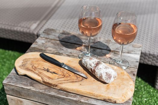 Two glasses of rose wine and a dried sausage with a knife on a wooden table in a garden on a sunny day