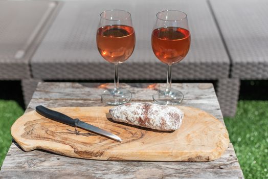 Two glasses of rose wine and a dried sausage with a knife on a wooden table in a garden on a sunny day