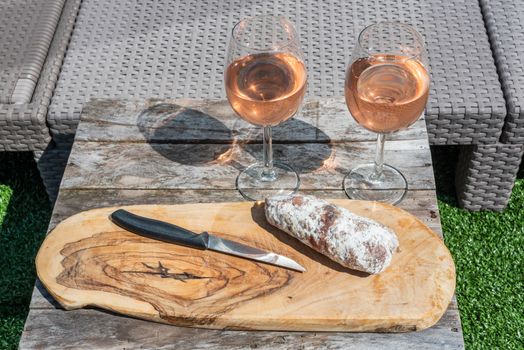 Two glasses of rose wine and a dried sausage with a knife on a wooden table in a garden on a sunny day