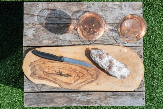 Two glasses of rose wine and a dried sausage with a knife on a wooden table in a garden on a sunny day