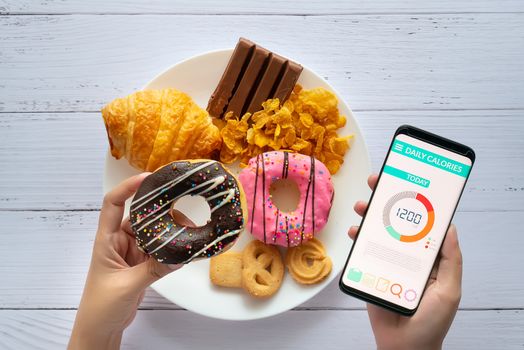 Calories counting and food control concept. woman using Calorie counter application on her smartphone with doughnut in hand and snack , cookies on plate at background