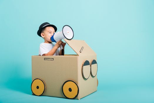 Happy Asian children boy smile in driving play car creative by a cardboard box imagination with megaphone, summer holiday travel concept, studio shot on blue background with copy space for text