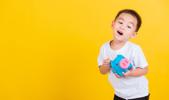 Asian Thai happy portrait cute little cheerful child boy smile putting coin money to the piggy bank and looking camera, studio shot isolated on yellow background with copy space