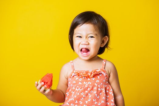 Happy portrait Asian baby or kid cute little girl attractive laugh smile wearing t-shirt playing holds cut watermelon fresh for eating, studio shot isolated on yellow background, healthy food and summer concept