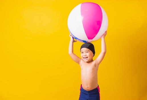 Summer vacation concept, Portrait Asian happy cute little child boy smiling in swimsuit hold beach ball, Kid having fun with inflatable ball in summer vacation, studio shot isolated yellow background