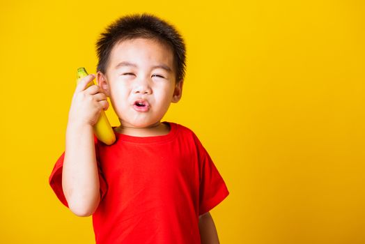 Happy portrait Asian child or kid cute little boy attractive smile wearing red t-shirt playing holds banana fruit pretending to be like a telephone, studio shot isolated on yellow background