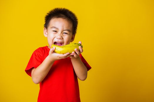 Happy portrait Asian child or kid cute little boy attractive smile wearing red t-shirt playing holds banana fruit, studio shot isolated on yellow background