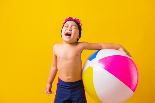 Summer vacation concept, Portrait Asian happy cute little child boy smiling in swimsuit hold beach ball, Kid having fun with inflatable ball in summer vacation, studio shot isolated yellow background