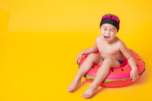 Summer vacation concept, Portrait Asian happy cute little child boy wear goggles, swimsuit hold beach watermelon inflatable ring, Kid have fun sit in inflatable, studio shot isolated yellow background
