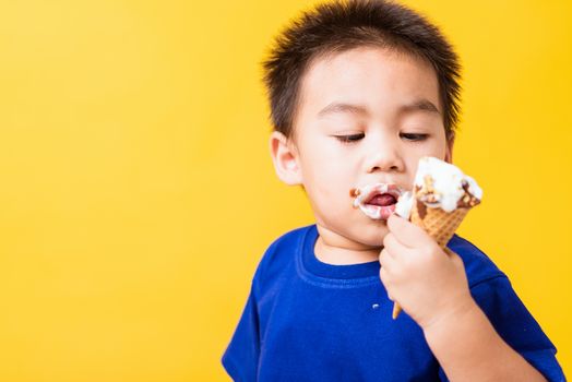 Happy portrait Asian child or kid cute little boy attractive laugh smile playing holds and eating sweet chocolate ice cream waffle cone, studio shot isolated on yellow background, summer concept