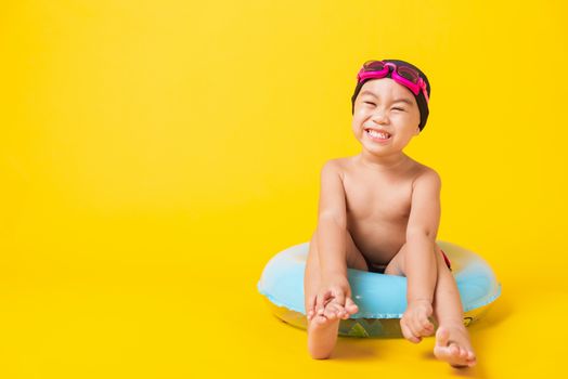 Summer vacation concept, Portrait Asian happy cute little child boy wear goggles, swimsuit hold beach blue inflatable ring, Kid have fun sit in inflatable, studio shot isolated yellow background