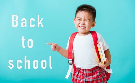 Back to school concept. Portrait Asian happy funny cute little child boy smile hug books and point finger to side away space, isolated blue background. Kid from preschool kindergarten with school bag