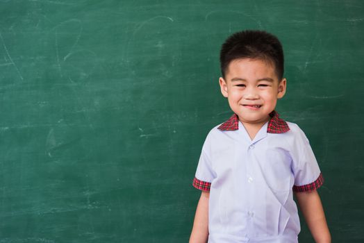 Back to School. Happy Asian funny cute little child boy from kindergarten in student uniform smiling on green school blackboard, First time to school education concept, studio shot