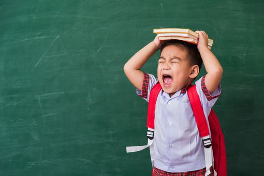 Back to School. Happy Asian funny cute little child boy from kindergarten in student uniform with school bag and book on head smiling on green school blackboard, First time to school education concept