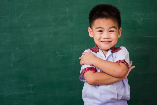 Back to School. Happy Asian funny cute little child boy from kindergarten in student uniform stand crossed arm smiling on green school blackboard, First time to school education concept, studio shot