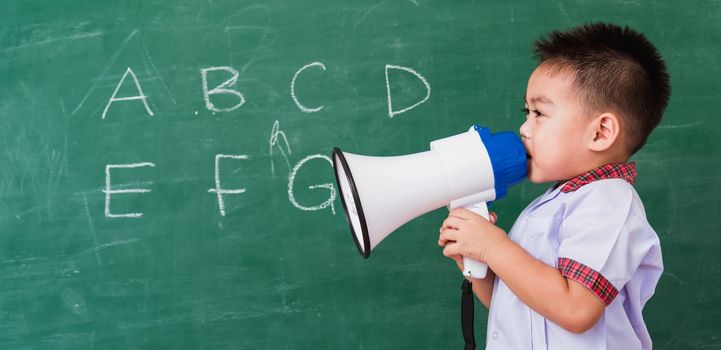 Back to School. Asian funny cute little child boy kindergarten preschool in student uniform speaking through megaphone against on green school blackboard, First time to school education concept