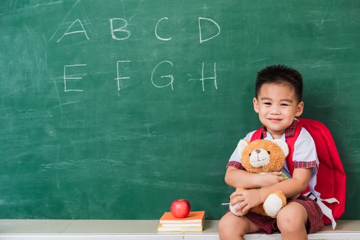 Back to School. Happy Asian funny cute little child boy from kindergarten in student uniform with school bag smiling and hugging teddy bear on green school blackboard, First time to school education