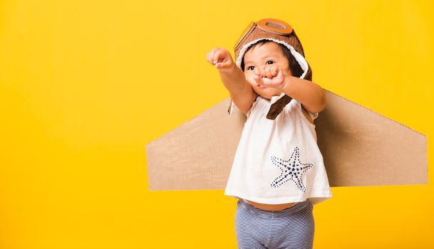 Happy Asian beautiful funny baby little girl smile wear pilot hat raise hand up play and goggles with toy cardboard airplane wings fly, studio shot isolated yellow background, Startup freedom concept