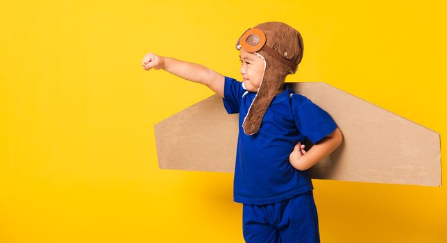 Happy Asian handsome funny child or kid little boy smile wear pilot hat play and goggles raise hand up with toy cardboard airplane wings flying, studio shot isolated yellow background, Startup freedom