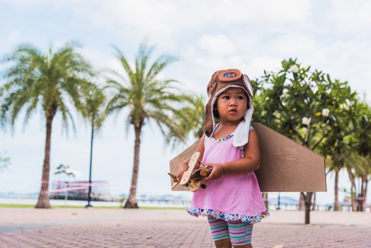 Happy Asian funny child or kid little girl smile wear pilot hat and goggles play toy cardboard airplane wing flying against summer sky cloud on trees garden background, Startup freedom concept