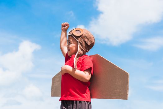 Happy Asian funny child or kid little boy smile wear pilot hat and goggles play toy cardboard airplane wing flying raises hand up against summer blue sky cloud background, Startup freedom concept