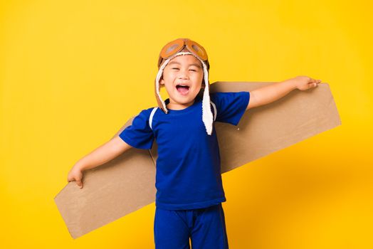 Happy Asian handsome funny child or kid little boy smile wear pilot hat playing and goggles with toy cardboard airplane wings flying, studio shot isolated yellow background, Startup freedom concept