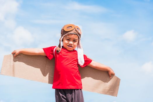 Happy Asian funny child or kid little boy smile wear pilot hat play and goggles with toy cardboard airplane wing flying outdoor against summer blue sky cloud background, Startup freedom concept