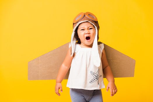 Happy Asian beautiful funny baby little girl smile wear pilot hat playing and goggles with toy cardboard airplane wings flying, studio shot isolated yellow background, Startup freedom concept