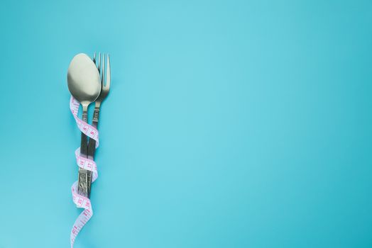 spoon and fork wrapped in measuring tape isolated on blue background with copy space. diet , food control and weight loss concept.