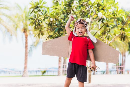 Happy Asian funny child or kid little boy smile wear pilot hat and goggles play toy cardboard airplane wing flying against summer sky cloud on trees garden background, Startup freedom concept