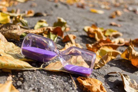 Hourglass as a symbol of the arrival of autumn time lie on the pavement surrounded by dry leaves