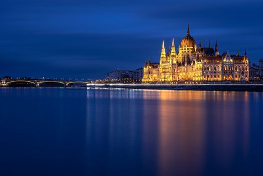 Hungarian Parliament Building built in the Gothic Revival style in Budapest viewed at night with Danube river and Margit bridge. Long exposure.