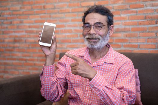 Happy senior man showing or presenting mobile phone application and pointing finger to smartphone on hand at home.
