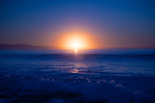 Sunset over ocean with waves crashing into shore with light lit in background