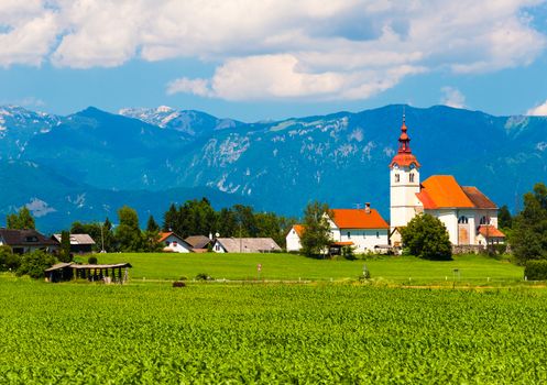 Old church in the field. Small village in Slovenia. Church building with orange-red roof and mountains on the background. Sunny summer day, green field and the mountain range.