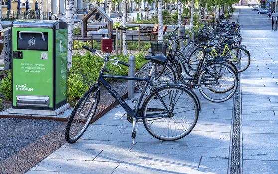 UMEA, SWEDEN - JUNE 10, 2020: A lot of bikes parked in long row on the street close to green recycling bin and small play park for children, central part of Umea city, Vasterbotten. Nice summer day