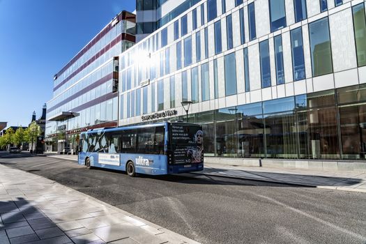 UMEA, SWEDEN - JUNE 10, 2020: Outdoor view of blue bus drive close to high modern hotel and shopping centrum building, Umea downtown. Early summer sun reflects in windows, morning. Umea, Vasterbotten