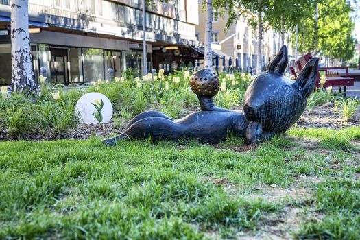 UMEA, SWEDEN - JUNE 10, 2020: Bronze sculpture of cat laying on back and playing with ball, closeup photo of brown kitten on green grass under birches, blurry yellow tulip flowers at background