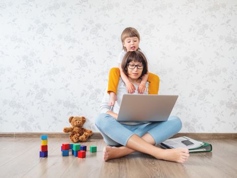 Mom and son at home. Mother works remotely with laptop, son sits on her neck. Freelance work in same time with raising children.