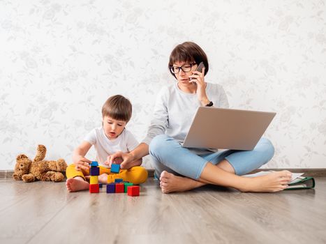 Mom and son at home quarantine because of coronavirus COVID19. Mother works remotely with laptop and smartphone, son plays with toy blocks. Freelance work.